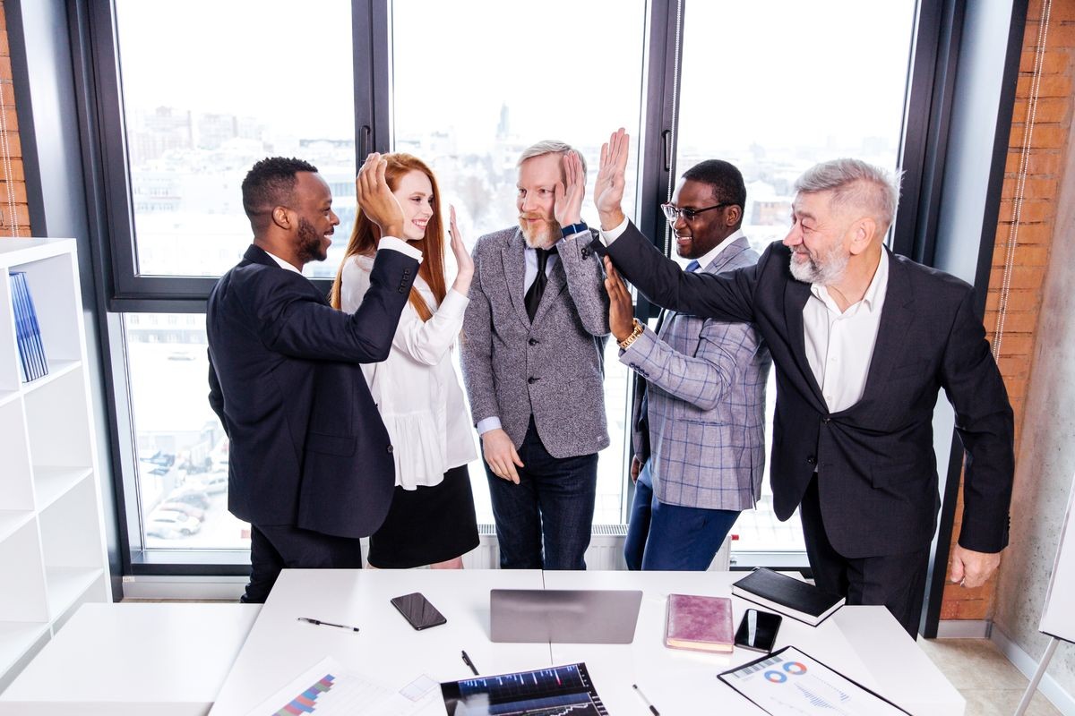 Business people of different ethnicity in a modern office are happy with the success of amicably giving five hands standing at the table with documents. Friendly joint work of business partners